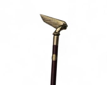 May include: A walking stick featuring a dark brown wooden shaft and a metallic gold-toned handle shaped like a stylized winged figure. The handle includes a small circular emblem on the upper shaft, and the base is finished with a metallic tip.