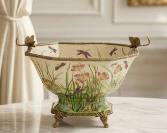 May include: A decorative ceramic bowl with a cream-colored base, featuring painted green grass, pink flowers, and purple dragonflies. The bowl has a gold-toned metal rim, ornate feet, and two small butterfly accents on the top edges.