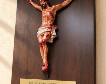 May include: A wall-mounted wooden crucifix featuring a detailed, hand-painted figure of Jesus with visible wounds, mounted on a dark brown wood plaque. A gold-toned metal plate at the bottom displays the text: FOR GOD SO LOVED THE WORLD THAT HE GAVE HIS ONE AND ONLY SON... JOHN 3:16.