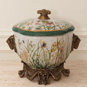 Puede incluir: Un tarro de cerámica con tapa y acabado craquelado, decorado con flores silvestres y mariposas coloridas. Presenta un borde verde oscuro, dos asas laterales doradas con forma de cabeza de carnero y una base metálica ornamentada.