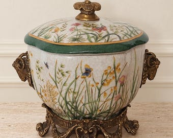 May include: A ceramic lidded jar with a crackle glaze finish, decorated with colorful wildflowers and butterflies. It features a dark green rim, two gold-toned ram head side handles, and a matching ornate gold-toned metal base and lid finial.