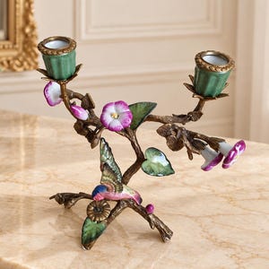 Puede incluir: Un candelabro doble con estructura de rama en tono bronce, decorado con soportes de cerámica verde para velas, flores moradas y blancas, hojas verdes y un pequeño pájaro con plumaje azul, rosa y verde en la rama.