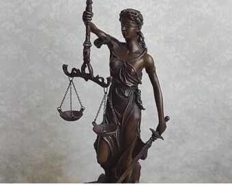 May include: A bronze-colored statue of Lady Justice featuring a blindfolded figure wearing a draped robe. The figure holds a raised pair of scales suspended by chains in the right hand and a downward-pointing sword in the left hand.
