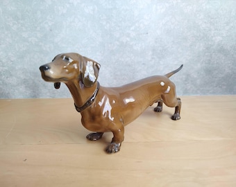 May include: A glossy ceramic figurine of a brown dachshund dog standing in a profile position. The dog has a smooth, light brown coat with darker brown shading on the ears, muzzle, and a thin collar around its neck, captured in a standing pose.