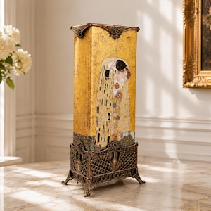 Può includere: Una lampada da tavolo alta e rettangolare con finitura dorata e una riproduzione del dipinto Il Bacio di Gustav Klimt sul pannello frontale. La lampada è sostenuta da una base in metallo scuro e decorata con motivo a griglia e quattro piedini.