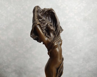May include: A bronze-toned sculpture of a figure with arms raised, holding a flowing fabric that drapes over the head and shoulders. The figure has a smooth, metallic finish and is depicted in a dynamic, upward-reaching pose with a serene expression.