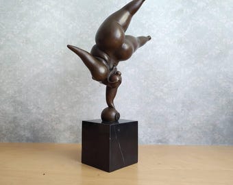 May include: A bronze-toned sculpture of a figure performing a handstand on a small sphere, which is mounted on a solid black square marble base. The figure is depicted in a dynamic pose with limbs extended, showcasing a smooth, metallic finish.