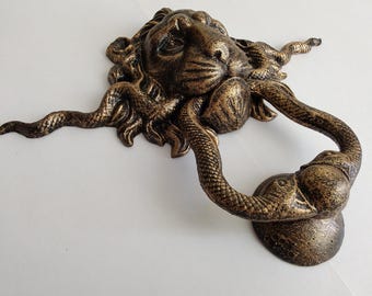 May include: A bronze-colored door knocker featuring a detailed lion head with a mane composed of intertwined snakes. The swinging part of the knocker is shaped like a snake, with its body forming a loop that connects to a circular base plate.