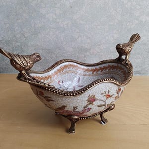 May include: An oval ceramic bowl with a white crackle glaze finish, featuring a colourful floral and bird pattern. The bowl is supported by three ornate bronze-toned metal feet and features two bronze-toned metal birds perched on the rim.