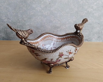 May include: An oval ceramic bowl with a white crackle glaze finish, featuring a colorful floral and bird pattern. The bowl is supported by three ornate bronze-toned metal feet and features two bronze-toned metal birds perched on the rim.