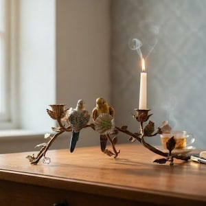 May include: A bronze-toned metal candle holder shaped like a curved branch with leaves and flowers. Two ceramic budgie figurines, one blue and white and one yellow, are perched on the branch. A white taper candle is lit, emitting thin wisps of smoke.