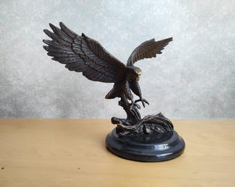 May include: A bronze-toned eagle sculpture with wings fully extended in a downward flight position. The bird is perched on a textured branch base, which is mounted on a smooth, circular black pedestal. The eagle features detailed feather textures.
