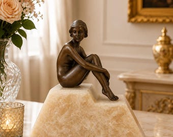 May include: A bronze-toned sculpture of a seated figure with short hair, positioned on a stepped, translucent, light-beige stone base. The figure is depicted in a resting pose with knees drawn toward the chest, showcasing a smooth, metallic finish.
