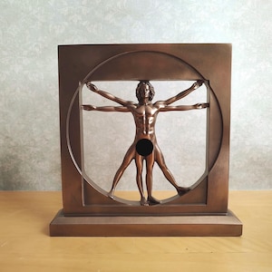 Pode incluir: Uma escultura de cor bronze representando o Homem Vitruviano, com uma figura humana central de braços e pernas estendidos dentro de uma moldura circular, inserida numa base e estrutura quadrada com um acabamento metálico liso.