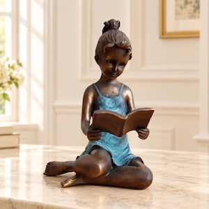 May include: A bronze-toned sculpture of a child sitting with legs bent, holding an open book. The figure has a dark metallic finish with a teal-coloured dress featuring a small floral detail at the neckline and hair pulled up in a bun.
