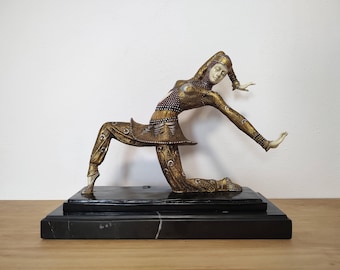 May include: A decorative figurine of a kneeling dancer in a gold and dark-toned outfit with intricate patterns, featuring a pale face and headpiece, mounted on a rectangular black marble base with white veining details.