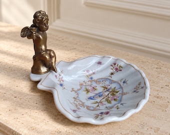May include: A white ceramic shell-shaped dish featuring a delicate floral and bird pattern in blue, pink, and green. A bronze-toned metal figurine of a seated cherub with wings is attached to the edge of the dish, serving as a decorative handle.