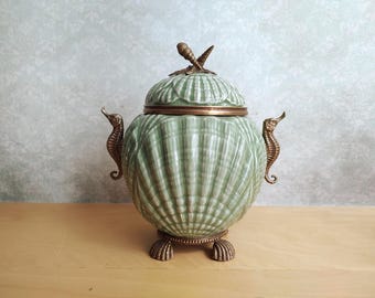 May include: A pale green ceramic lidded jar shaped like a seashell, featuring brass-toned metal accents including two seahorse-shaped side handles, three shell-shaped feet, and a decorative finial on top consisting of two crossed shells.