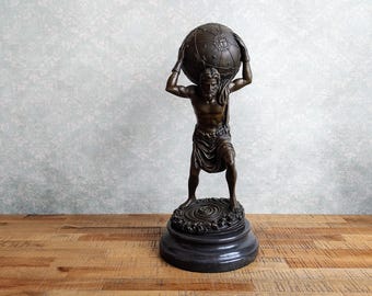 May include: A bronze-toned sculpture of a muscular figure standing on a circular base, holding a large celestial globe above their head. The figure wears a draped cloth around the waist, and the globe features engraved star and sun patterns.