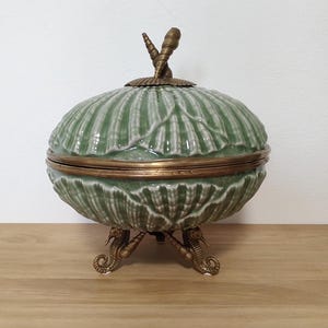 Może przedstawiać: Ceramiczna miska z pokrywką, posiadająca szałwiowo-zielony wzór muszli na zewnątrz oraz mosiężną metalową krawędź. Pokrywka zwieńczona jest uchwytem w kształcie muszli, a cała miska wspiera się na trzech metalowych nóżkach w kształcie koników morskich.