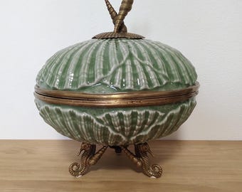 May include: A lidded ceramic bowl featuring a sage green shell-patterned exterior with a brass-toned metal rim. The lid is topped with a brass-toned shell finial, and the bowl is supported by three brass-toned metal legs shaped like seahorses.