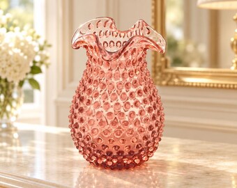 May include: A rose-colored glass vase featuring a hobnail texture across its rounded body and a ruffled, scalloped rim. The translucent vessel displays a consistent pink hue throughout its surface, highlighting the raised circular pattern.