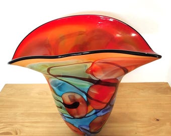 May include: A hand-blown glass vase with a wide, flared rim. The body features a vibrant swirl of colors including bright red, orange, and light blue, accented by dark brown linear patterns that weave through the translucent glass surface.