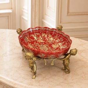 Puede incluir: Un cuenco de cerámica roja con borde festoneado y complejos patrones florales dorados, sostenido por tres figuras de querubines color latón. Las figuras están conectadas por un detalle de cuerda decorativa en la base del cuenco.