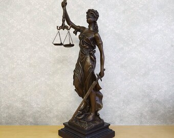 May include: A bronze-colored statue of a blindfolded figure standing on a square base. The figure holds a set of scales in the right hand and a sword in the left hand, wearing draped clothing that flows down to the feet on a dark pedestal.