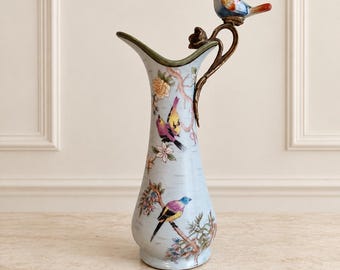 May include: A light blue ceramic vase featuring a painted design of colorful birds and floral branches. The vase has a curved rim and a bronze-toned metal handle that extends upward, topped with a small, three-dimensional ceramic bird figure.