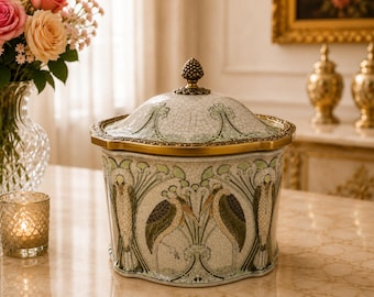 May include: A ceramic lidded jar featuring a crackled white glaze with green and brown illustrations of long-legged birds and stylized floral motifs. The jar has a gold-toned metal rim and a decorative finial shaped like a pinecone.