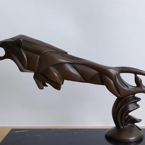 Può includere: Una scultura color bronzo di un leone che balza con un design geometrico e sfaccettato. Il leone è raffigurato in posizione di salto con la bocca aperta che mostra i denti. La figura è sostenuta da una base curva e poggia su un basamento nero rettangolare.
