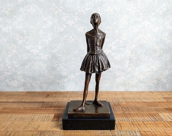 May include: A bronze-toned sculpture of a standing ballerina wearing a fitted bodice and a pleated tutu. The figure is positioned with hands behind the back, head tilted upward, and is mounted on a square base atop a black rectangular plinth.