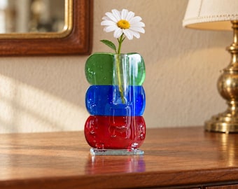 May include: A clear glass vase with three stacked, rounded sections colored green, blue, and red. The vase features a single central opening holding a white daisy with a yellow center, showcasing a smooth, translucent finish throughout.