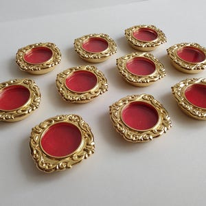 May include: A collection of ten small, circular gold-toned metal frames with ornate, scroll-like edges. Each frame features a recessed, vibrant red circular centre. The pieces are arranged in a scattered pattern, showcasing the metallic finish.
