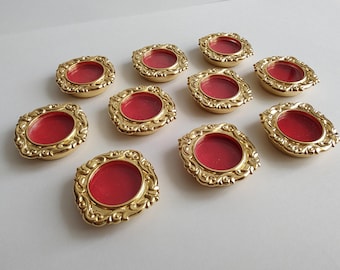 May include: A collection of ten small, circular gold-toned metal frames with ornate, scroll-like edges. Each frame features a recessed, vibrant red circular center. The pieces are arranged in a scattered pattern, showcasing the metallic finish.