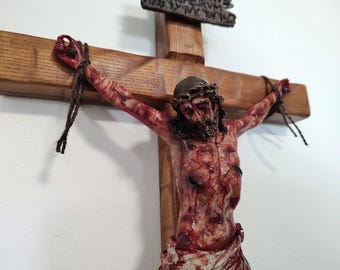 May include: A wooden crucifix featuring a detailed, hand-painted figure of a person with a crown of thorns, depicted with visible wounds and blood. The figure is attached to the cross with dark twine, and a small sign with text is at the top.