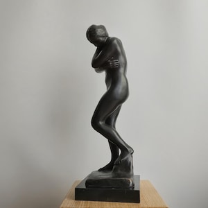 May include: A dark charcoal-coloured sculpture of a standing figure with arms crossed over the chest. The figure is depicted in a slightly hunched posture, with the head tilted downward, mounted on a rectangular black base with a smooth finish.
