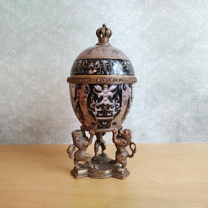 Op de afbeelding: Een decoratieve eivormige houder met een kroonvormige knop, voorzien van een zwart en roze oppervlak met gedetailleerde heraldische illustraties. Het object rust op een bronskleurige metalen voet bestaande uit drie gebeeldhouwde leeuwen.