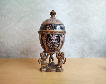 May include: A decorative egg-shaped container with a crown-shaped finial, featuring a black and pink surface with intricate heraldic illustrations. The object is supported by a bronze-toned metal base consisting of three sculpted lion figures.