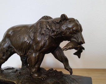 May include: A bronze-colored sculpture of a bear walking on a textured base. The bear is depicted with detailed fur texture and is holding a fish in its mouth. The sculpture is finished in a dark metallic brown tone with visible highlights on the fur.