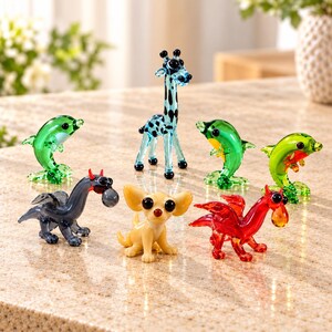 Könnte beinhalten: Eine Sammlung von sieben kleinen Tierfiguren aus Glas, darunter eine hellblaue Giraffe mit schwarzen Flecken, drei grüne Delfine, ein dunkelgrauer Drache mit roten Hörnern, ein roter Drache mit Flügeln und ein beigefarbener Hund mit großen schwarzen Augen.