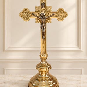 Op de afbeelding: Een goudkleurig metalen crucifix op een ronde voet. Het kruis heeft versierde, klaverbladvormige uiteinden en een centrale zilverkleurige figuur van Jezus. Boven de figuur bevindt zich een klein plaatje met de letters INRI in reliëf.