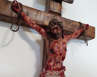 May include: A wooden crucifix featuring a detailed figure of a person with a crown of thorns, secured to the cross with dark brown rope. The figure displays painted red markings across the body, arms, and face, suggesting a wounded appearance.