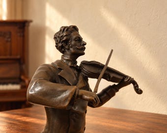 May include: A bronze-toned sculpture of a musician with a mustache, wearing a formal jacket and bow tie, captured in a side profile while playing a violin with a bow held in the right hand and the instrument positioned on the left shoulder.
