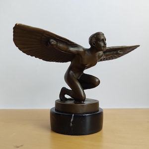 Könnte beinhalten: Eine bronzefarbene Skulptur einer knienden Figur mit ausgebreiteten Flügeln in einer nach vorne geneigten Haltung. Die Figur ist auf einem zylindrischen Sockel aus schwarzem Marmor mit feiner weißer Maserung montiert, was für einen stabilen Stand sorgt.