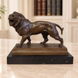 Könnte beinhalten: Eine bronzefarbene Skulptur eines stehenden Löwen mit geöffnetem Maul, detaillierten Gesichtszügen und muskulösem Körperbau. Der Löwe ist auf einem rechteckigen Bronzesockel montiert, der auf einem massiven, glatten schwarzen Marmorsockel steht.