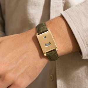May include: A rectangular gold-tone watch with a brushed metal finish and a dark olive green leather strap featuring a crocodile texture. The watch face displays a digital-style hour window at the top and a curved minute indicator window below.