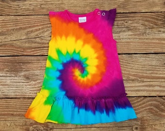 May include: A colorful tie-dye dress with a ruffled hem. The dress is sleeveless and has a round neckline. The colors are pink, orange, yellow, green, blue, and purple.