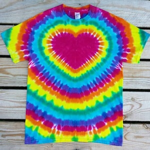 May include: A vibrant tie-dye t-shirt featuring a large heart design in the center. The shirt showcases a spectrum of colors, including pink, yellow, orange, blue, and purple, radiating outwards from the heart shape. The short-sleeved tee is made of soft fabric.